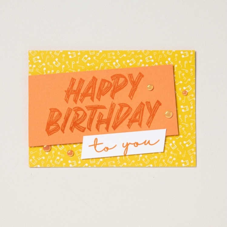 Handmade birthday card featuring an orange stamped “Happy Birthday” design layered on patterned paper.