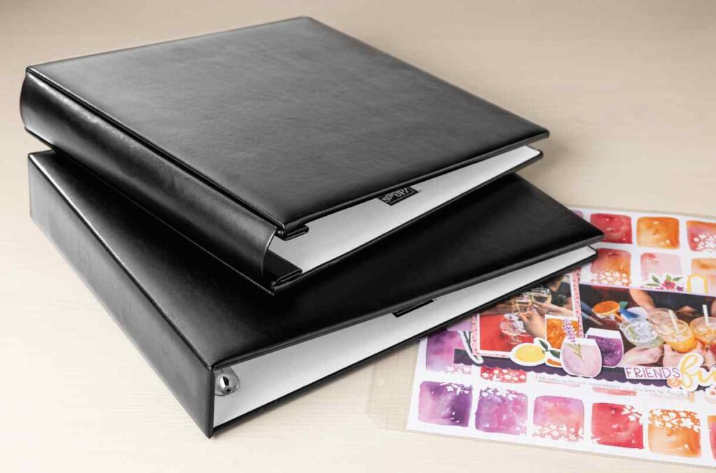 Gestapelde zwarte scrapbookalbums met D-ringen en bedrukte lay-outs ernaast, die platte foto- en paginaopslag demonstreren voor georganiseerde scrapbooking.
