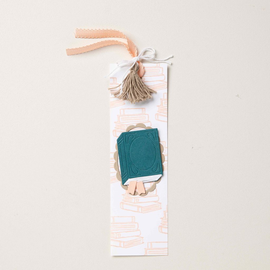 Marque-page fait main avec un ornement de livre sarcelle, un pompon, un ruban et un fond subtil à motif de livre.
