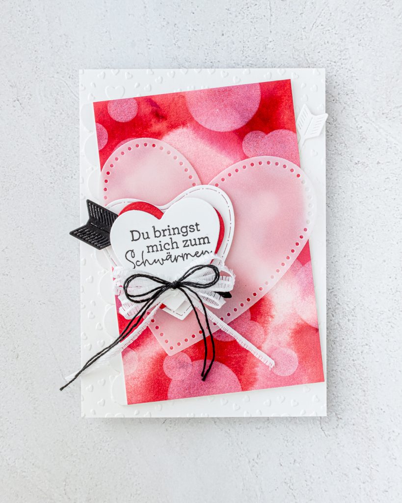 Carte artisanale finie, présentant un fond bokeh rose vif, des poinçons en forme de cœur superposés, des rubans décoratifs et des embellissements.