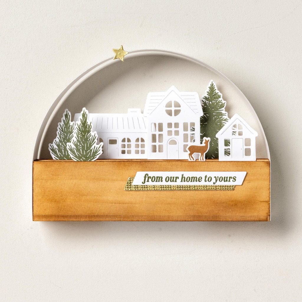 Kerstdisplay in boogvorm met witte papieren huisjes, groene bomen, een hert, een gouden ster en de tekst 'van ons huis naar het jouwe'.