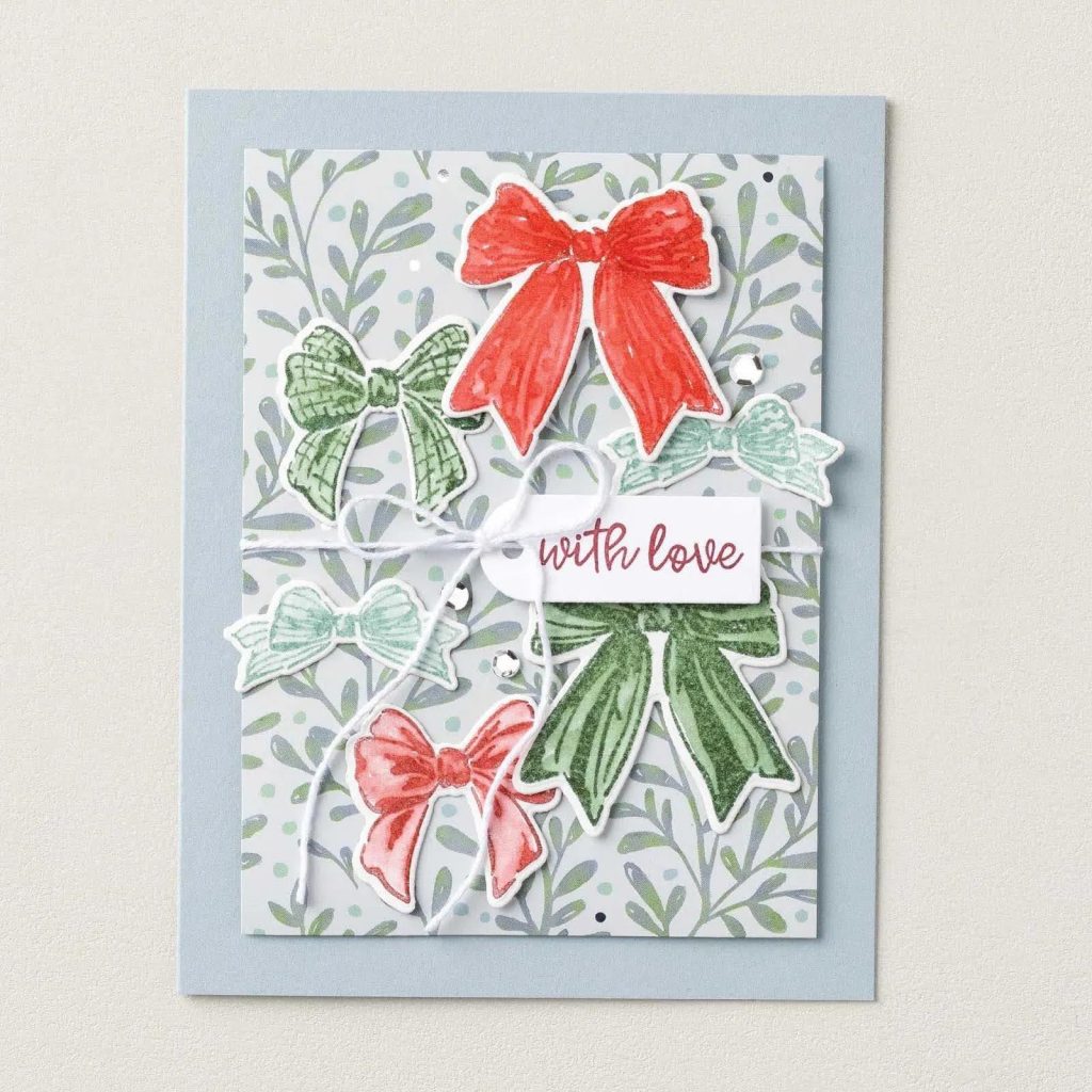 Pale blue card with red, green, and mint stamped bows on leafy paper, white twine, and a small “with love” tag.