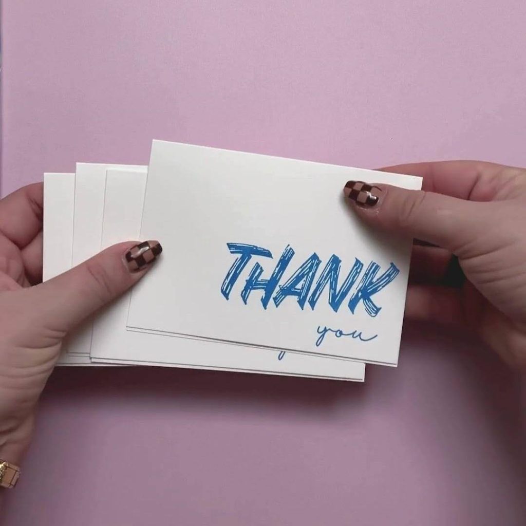 Deux mains tiennent une pile de cartes blanches sur lesquelles figurent, sur fond rose, le mot « THANK » en bleu vif et le mot « you » en lettres cursives.