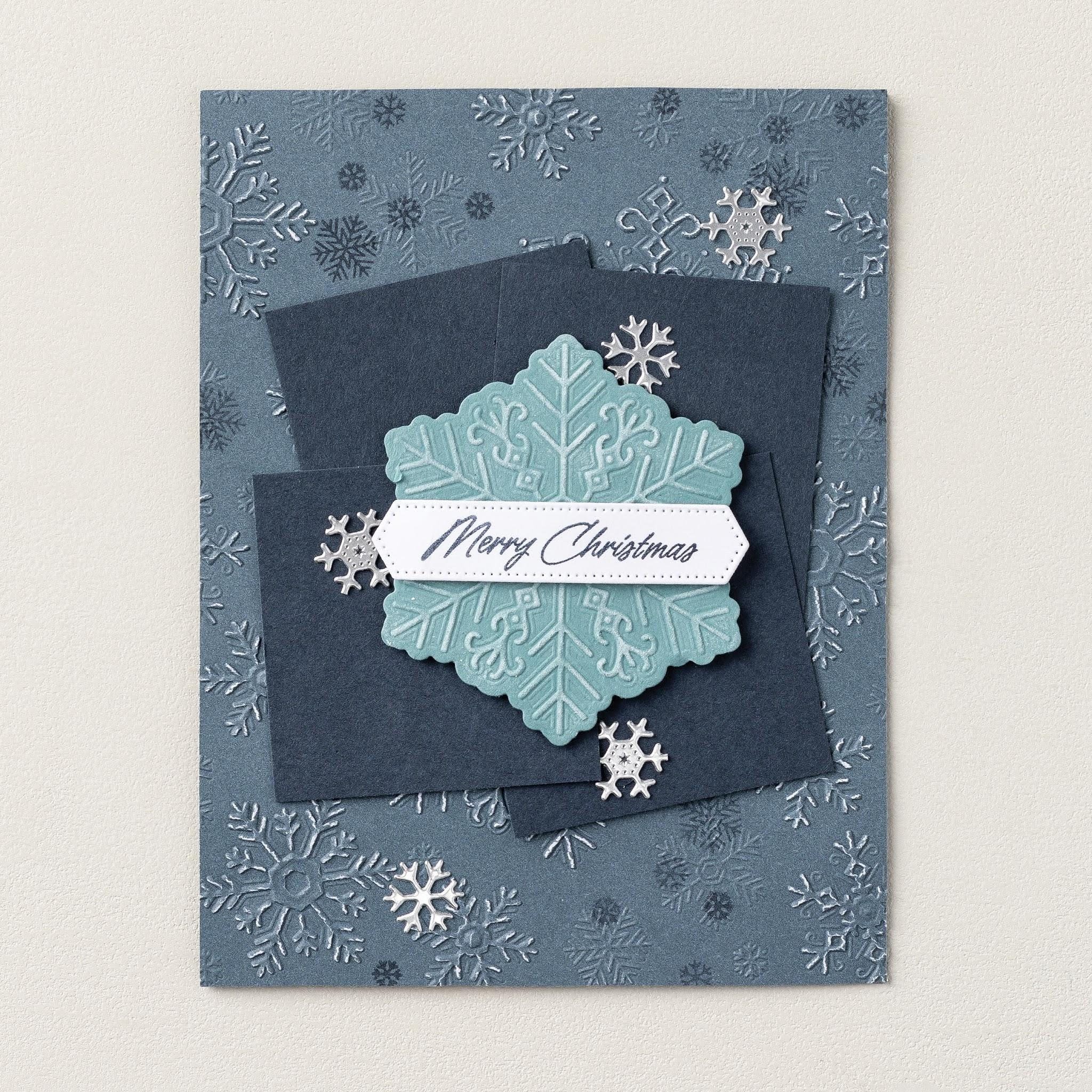 Carton bleu avec fond de flocon de neige embossé, panneaux bleu marine qui se chevauchent, un grand flocon aqua, flocons de neige argentés et une étiquette « Joyeux Noël ».
