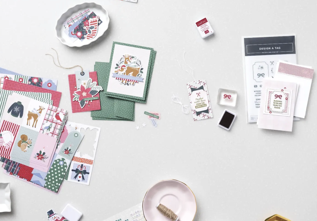 Du matériel pour la création de cartes de Noël disposé sur une table, comprenant notamment du papier à motifs, des cartes faites main, des étiquettes-cadeaux, des tampons et des tampons encreurs.