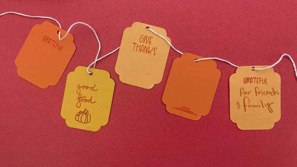 Des étiquettes de remerciement pour Thanksgiving, faites à la main, de couleur orange et jaune, avec des messages créés à l’aide de tampons, suspendues à de la ficelle blanche