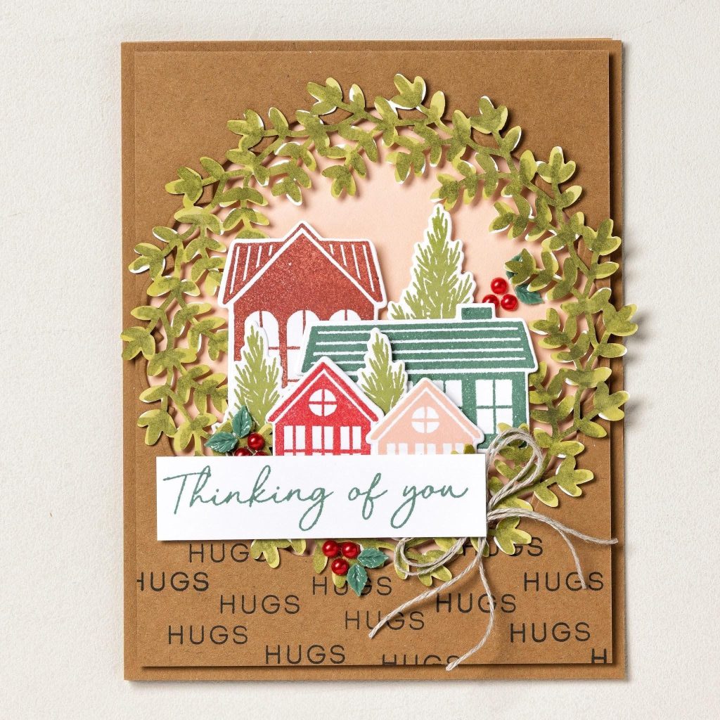 Carte marron ornée de maisons colorées au sein d'une couronne de feuillage, de baies rouges, d'un nœud en ficelle et d'une inscription « Je pense à vous » au-dessus du mot « HUGS » créé.