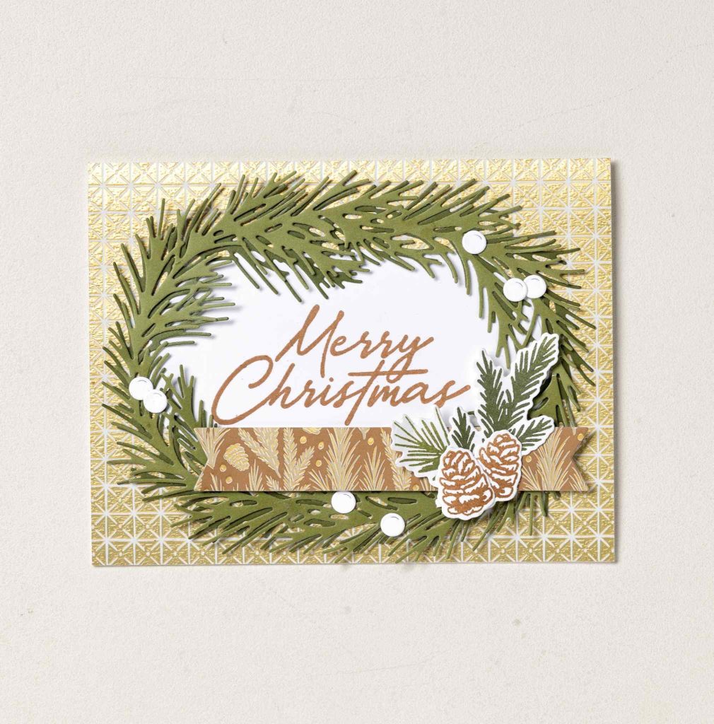 Christmas card with a green evergreen wreath, pinecones, white berry accents, a gold patterned background, and a brown “Merry Christmas” greeting.