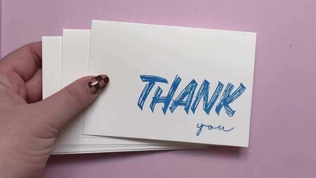 A hand holds a stack of white note cards with a large blue “THANK” and script “you” on a pink surface.
