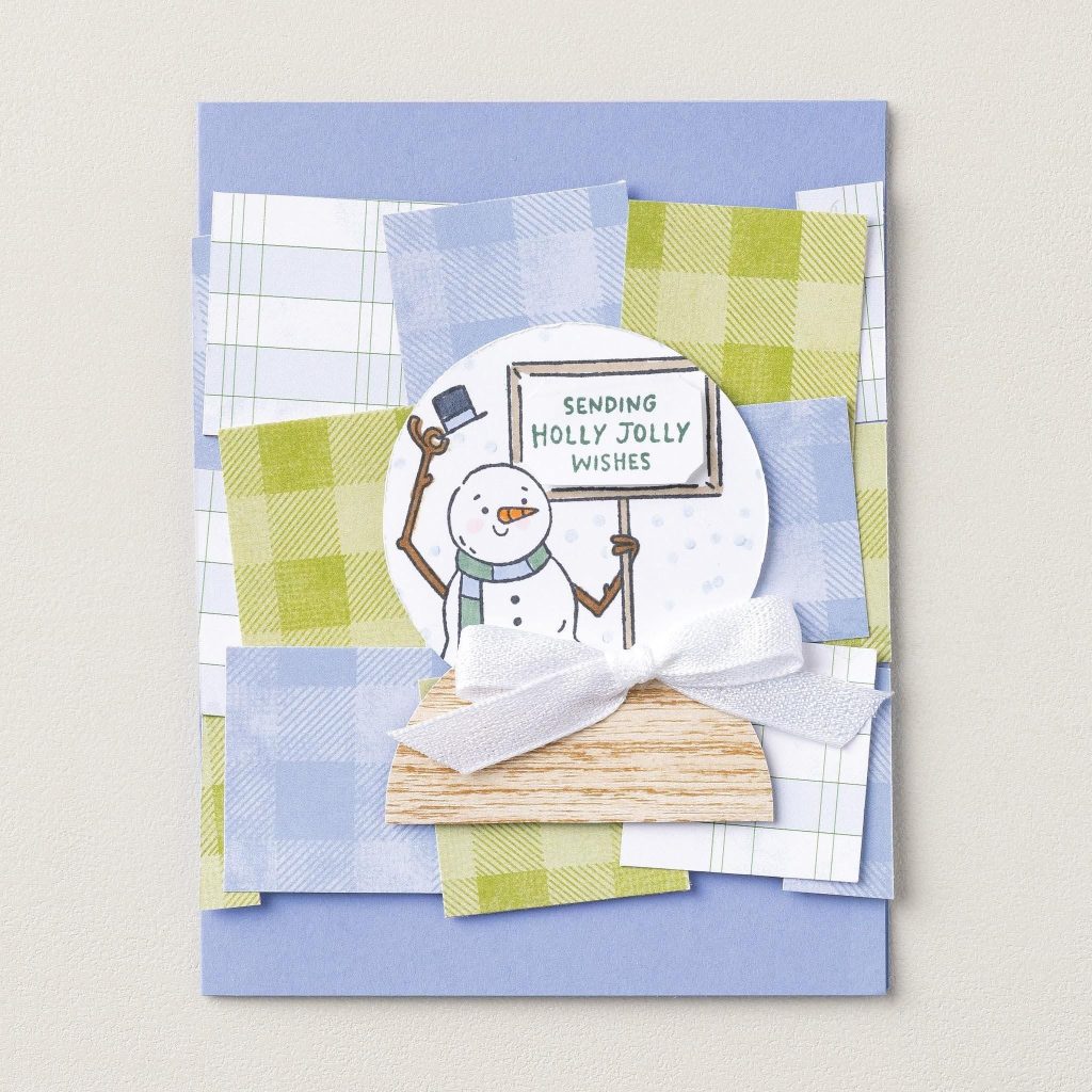 Un bonhomme de neige tient une pancarte « Envoyer des vœux joyeux à Holly » à l’intérieur d’une boule à neige sur des couches à carreaux bleus et verts avec un nœud blanc.